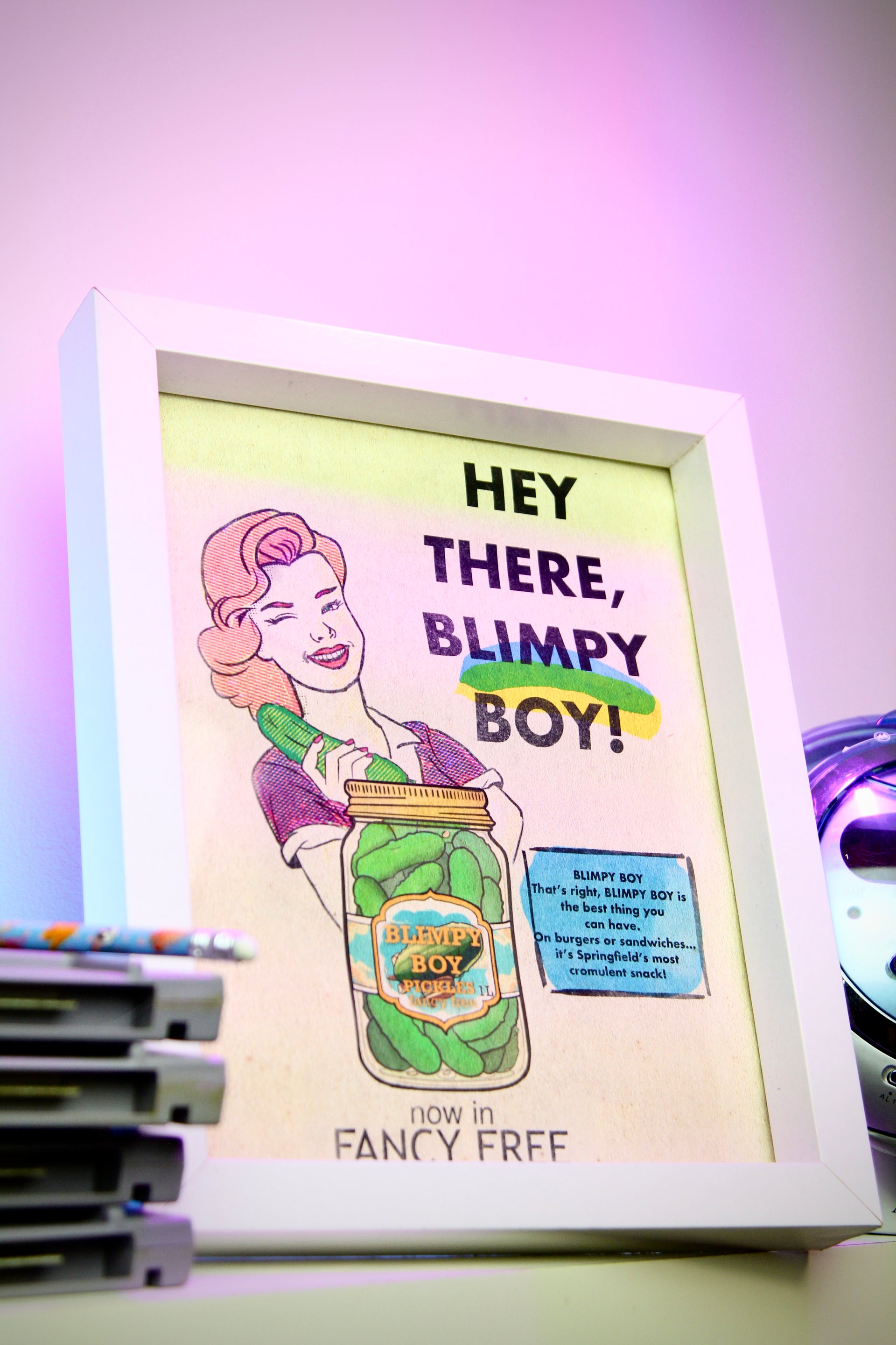 Framed artwork with a vintage-style illustration of a woman holding a jar labeled 'Blimpy Boy' on a shelf.