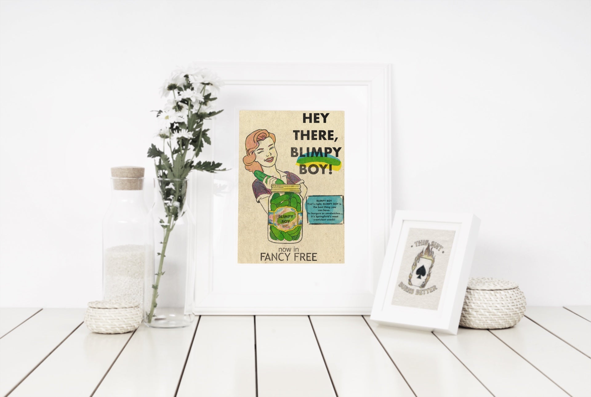 Vintage-style illustration of a woman winking at a jar of pickles labeled "Blimpy Boy Pickles," with accompanying text about them being Springfield's most cromulent snack on table next to ornaments and a smaller frame to the right.