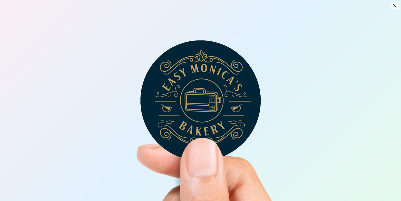 Woman holding a dark blue circular vinyl sticker with a vintage-style gold logo for "EASY MONICA'S BAKERY" featuring ornate scrolls and a central illustration of a microwave oven.
