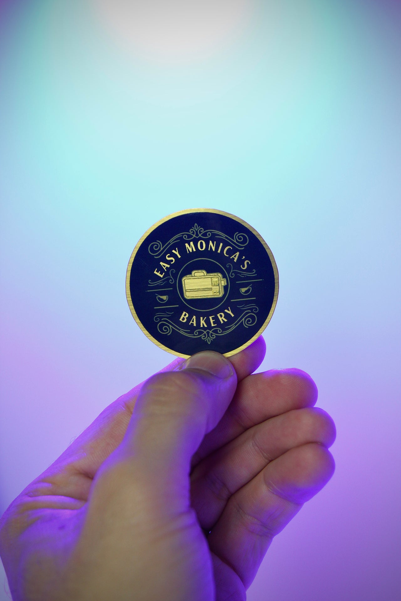 Hand holding a round blue and gold card with 'Easy Monica's Bakery' branding against a gradient background.