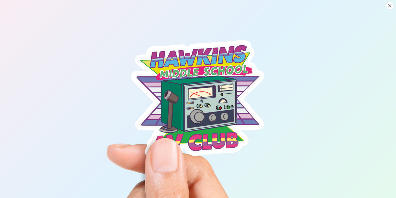 Woman's hand holding Vibrant retro-style vinyl sticker with "HAWKINS MIDDLE SCHOOL AV CLUB" text above and below an illustration of a vintage green radio/intercom, all on a neon pink and green background.