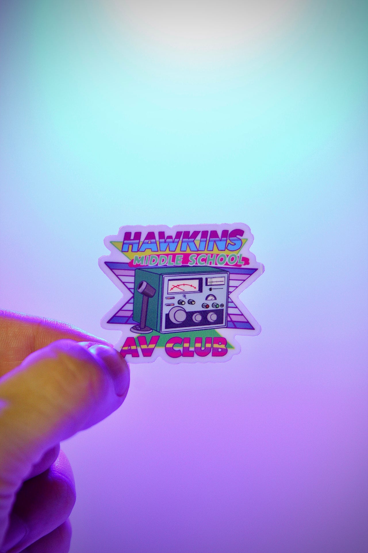 Sticker of Hawkins Middle School AV Club held by a hand against a gradient background