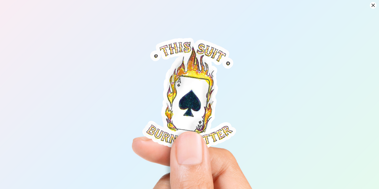 Hand holding a a vibrant vinyl sticker featuring a white Ace of Spades playing card engulfed in yellow and orange flames, with the text "THIS SUIT" above and "BURNS BETTER" below, on a colorful abstract background